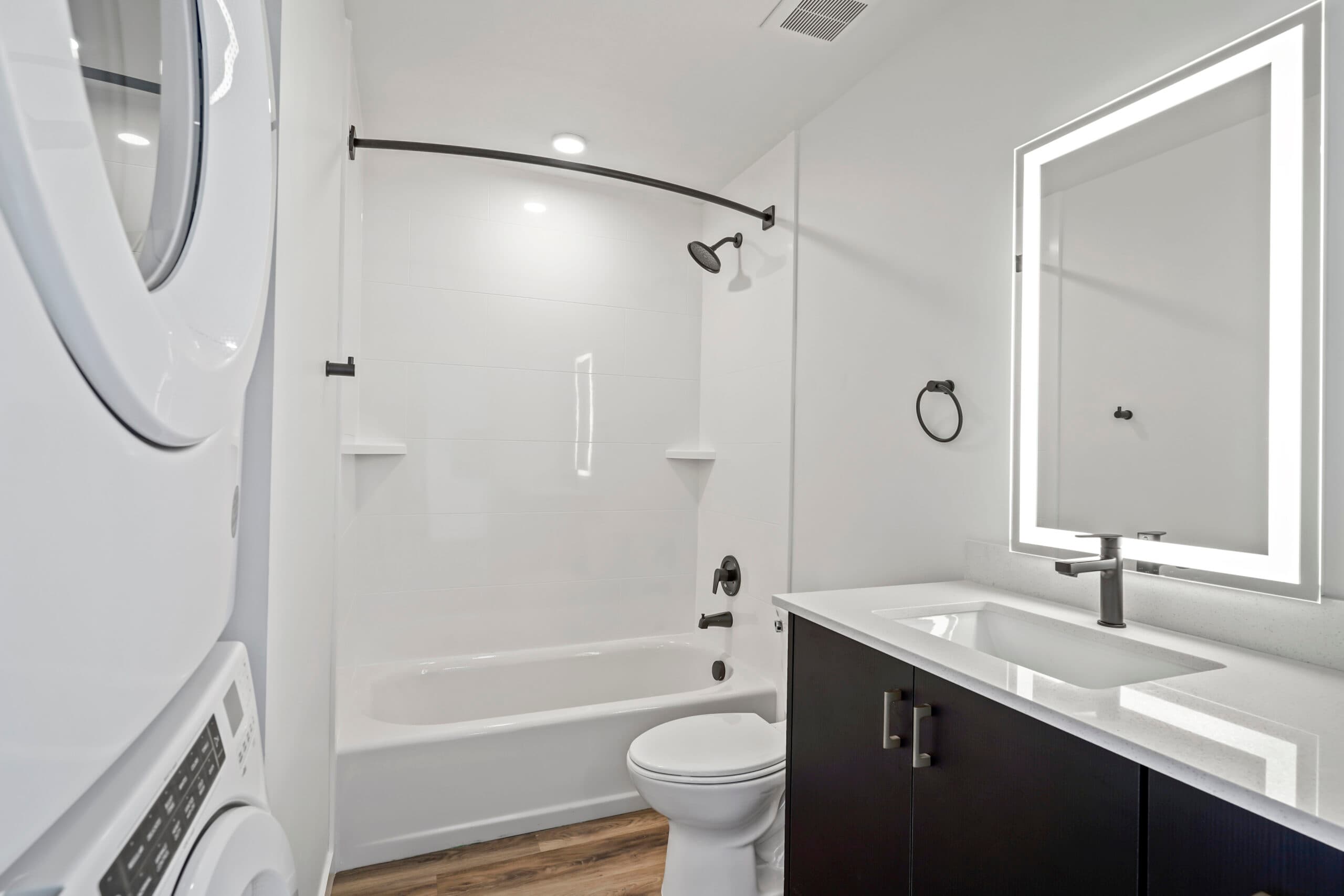 Modern bathroom at 343 Apartments Salt Lake City with stacked washer-dryer, bathtub-shower combo with black fixtures, a toilet, vanity featuring lighted mirror and black cabinet, all accented by clean white walls and wood flooring.