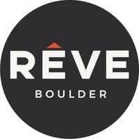 Logo for RÊVE Boulder Apartments. The word RÊVE is in large white letters with an orange triangle over the E, and BOULDER is written below in smaller white letters, all on a dark circular background—perfect for Boulder apartment branding.