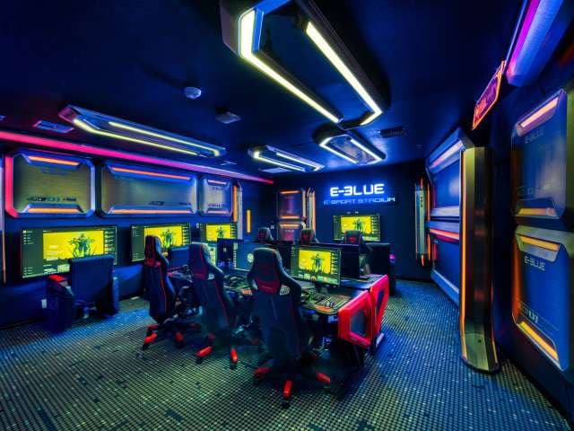 A futuristic gaming room with neon lighting, featuring multiple high-end gaming computers, large monitors, and ergonomic chairs arranged in a row. E-Blue Esport Stadium is illuminated on the back wall.
