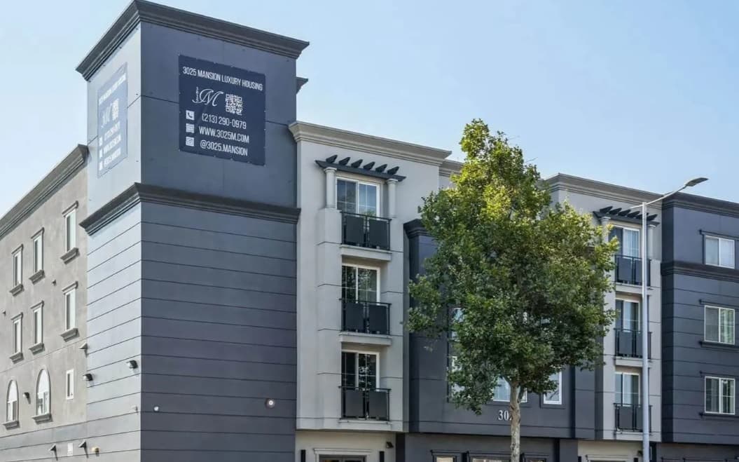 A modern gray and white apartment building—3025 Mansion—features small balconies, large windows, and a tree in front. The building displays a sign with contact information for luxury housing.