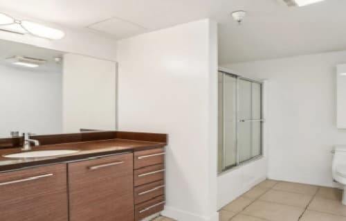 A modern bathroom in the 3025 Mansion features a wooden vanity, large mirror, sleek sink, glass-enclosed shower, and toilet. The space is accented by white walls and beige tiled flooring for a clean, luxurious feel.