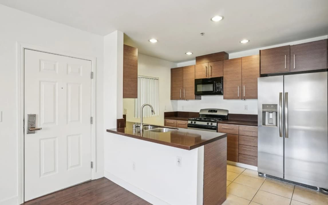 Modern kitchen in the 3025 Mansion with brown cabinets, stainless steel appliances including a refrigerator and gas stove, built-in microwave, double sink with brown countertop, and white walls with recessed lighting. White entry door on the left.