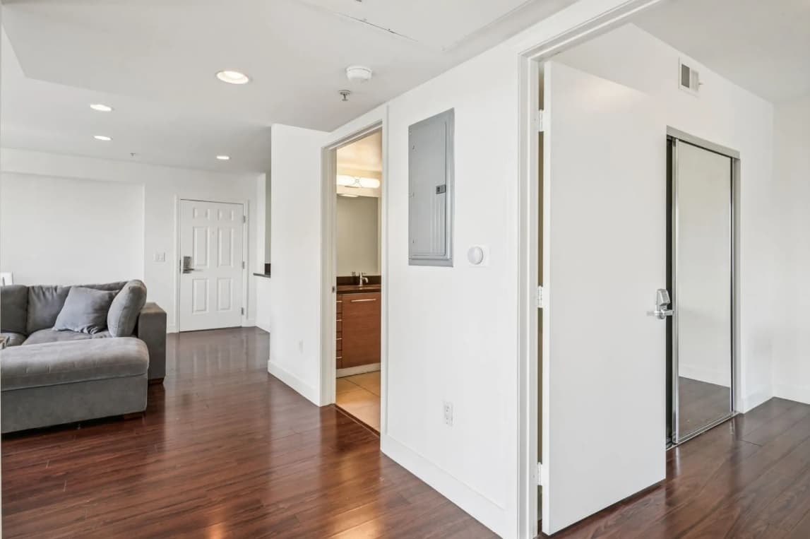 A modern 3025 Mansion apartment interior with wood floors, white walls, an open door to a bathroom with a vanity, a gray sectional sofa, and a mirrored closet door.