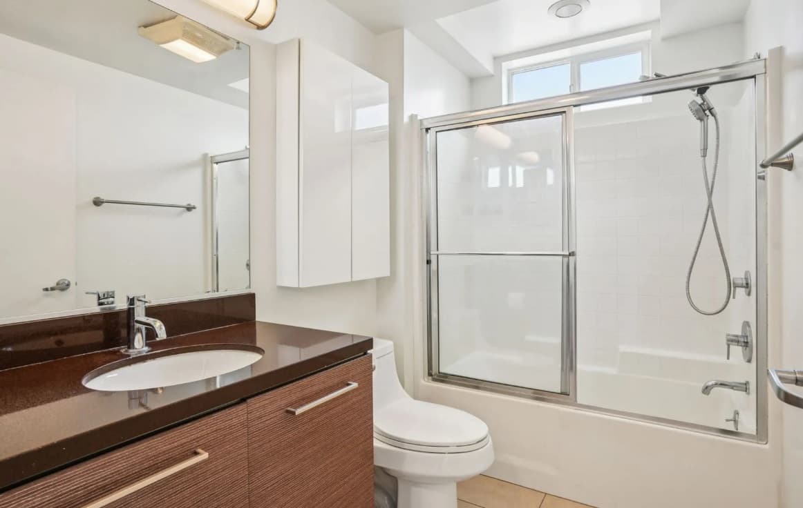 Modern bathroom in the 3025 Mansion featuring a brown vanity, white sink and toilet. Enjoy a glass-enclosed shower and bathtub, white tile walls, a large mirror, and bright natural light from a window above the shower.