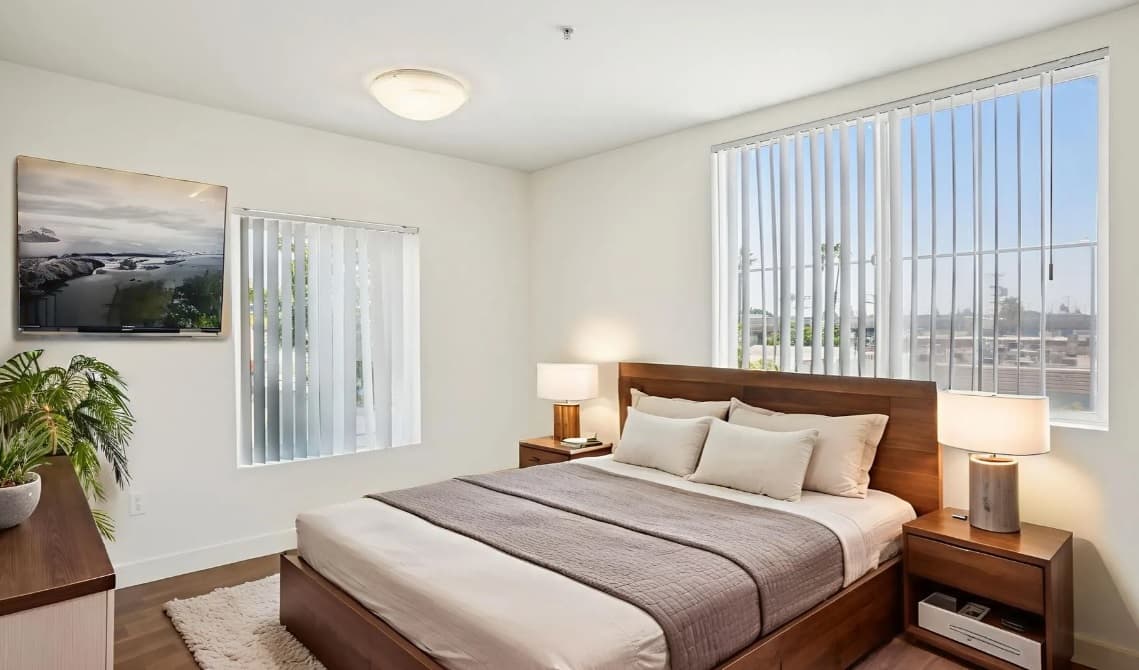 A bright, modern bedroom in the 3025 Mansion features a wooden bed, neutral bedding, two nightstands with lamps, large windows with vertical blinds, a potted plant, and a wall-mounted TV displaying a nature scene.