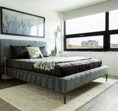 Modern bedroom at 3rd Street Flats Reno with a large bed featuring gray bedding, decorative pillows, and a light throw. A vase with flowers and framed photo rest on bedside tables. Large windows fill the space with natural light and city views.