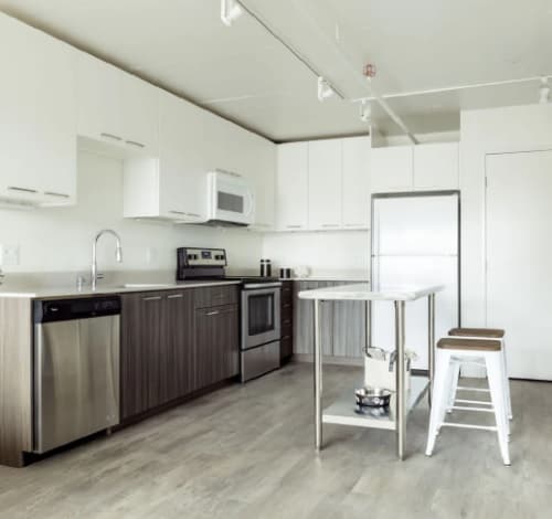 Modern kitchen at 3rd Street Flats Reno with white upper cabinets, wood lower cabinets, stainless steel appliances, a small island with shelves, and a white stool on light wood flooring. The space is bright and minimalistic.