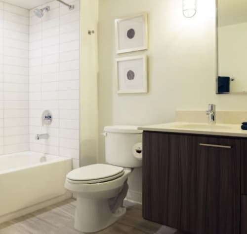 Modern bathroom at 3rd Street Flats Reno with a white bathtub and shower, toilet, dark wood vanity with sink, large mirror, and two minimalist framed artworks on a light-colored wall. The floor is light-tiled.