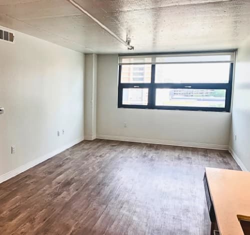 A bright, empty room at 3rd Street Flats Reno features light-colored walls, wood-style flooring, a large window welcoming natural light, and a small part of a countertop visible in the lower right corner.