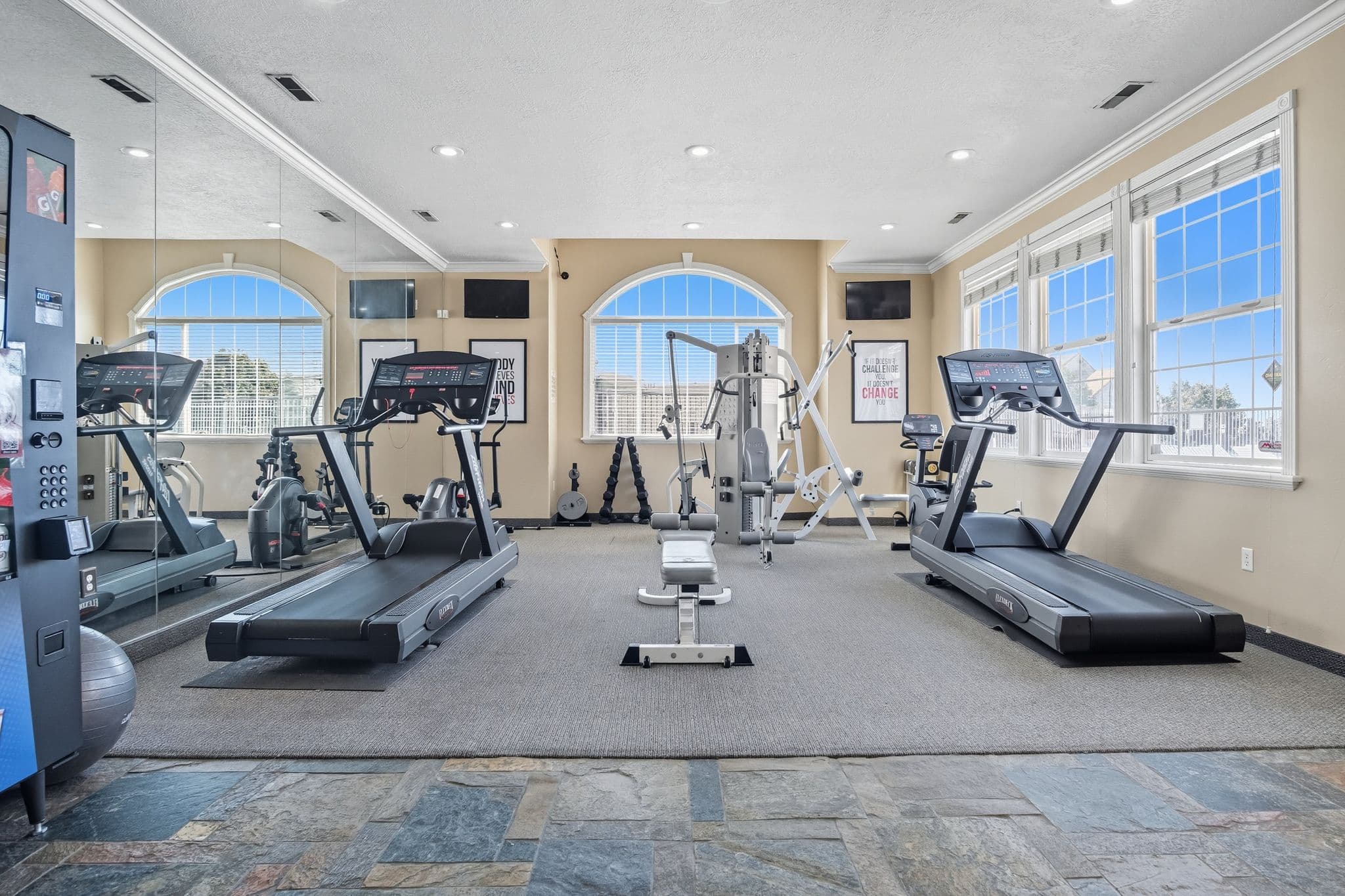 A bright, modern gym at Ventana Student Housing Orem features two treadmills, a multi-function weight machine, free weights, large mirrors, and windows letting in lots of natural light. The floor is covered with slate tiles and gray carpet.