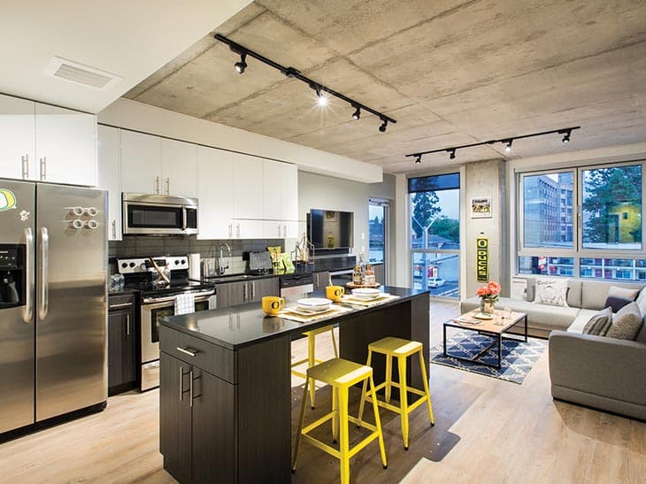 Modern apartment kitchen and living area at Theory U District, featuring stainless steel appliances, white upper cabinets, a dark island with two yellow stools, and a cozy gray sofa near large windows filling this student housing with natural light.