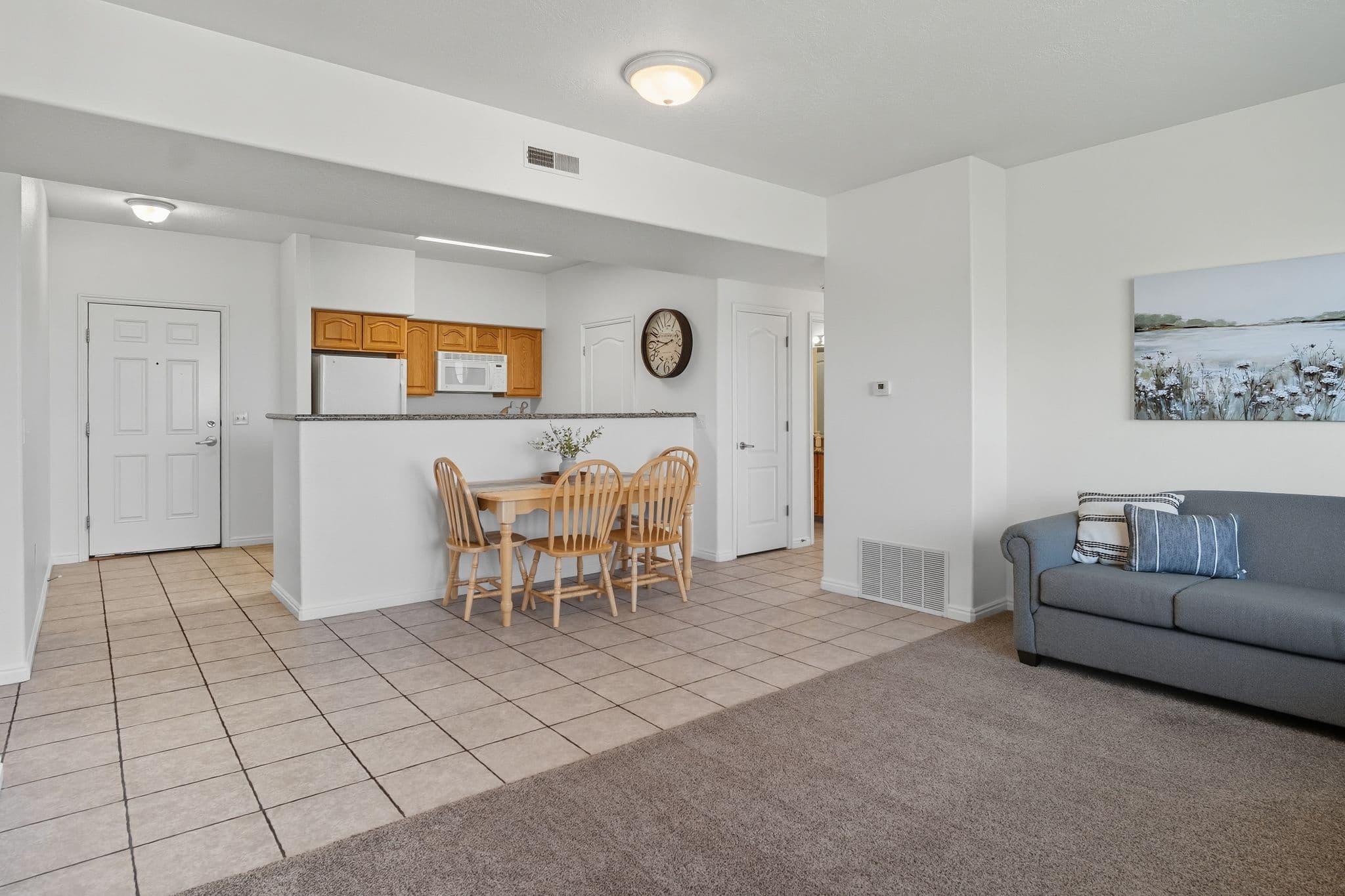 Open-plan living area at Ventana Student Housing Orem with tiled kitchen and dining space, light wood dining table with four chairs, white cabinets, a microwave, and a gray sofa with patterned pillows on carpeted floor. Walls are white and feature art.
