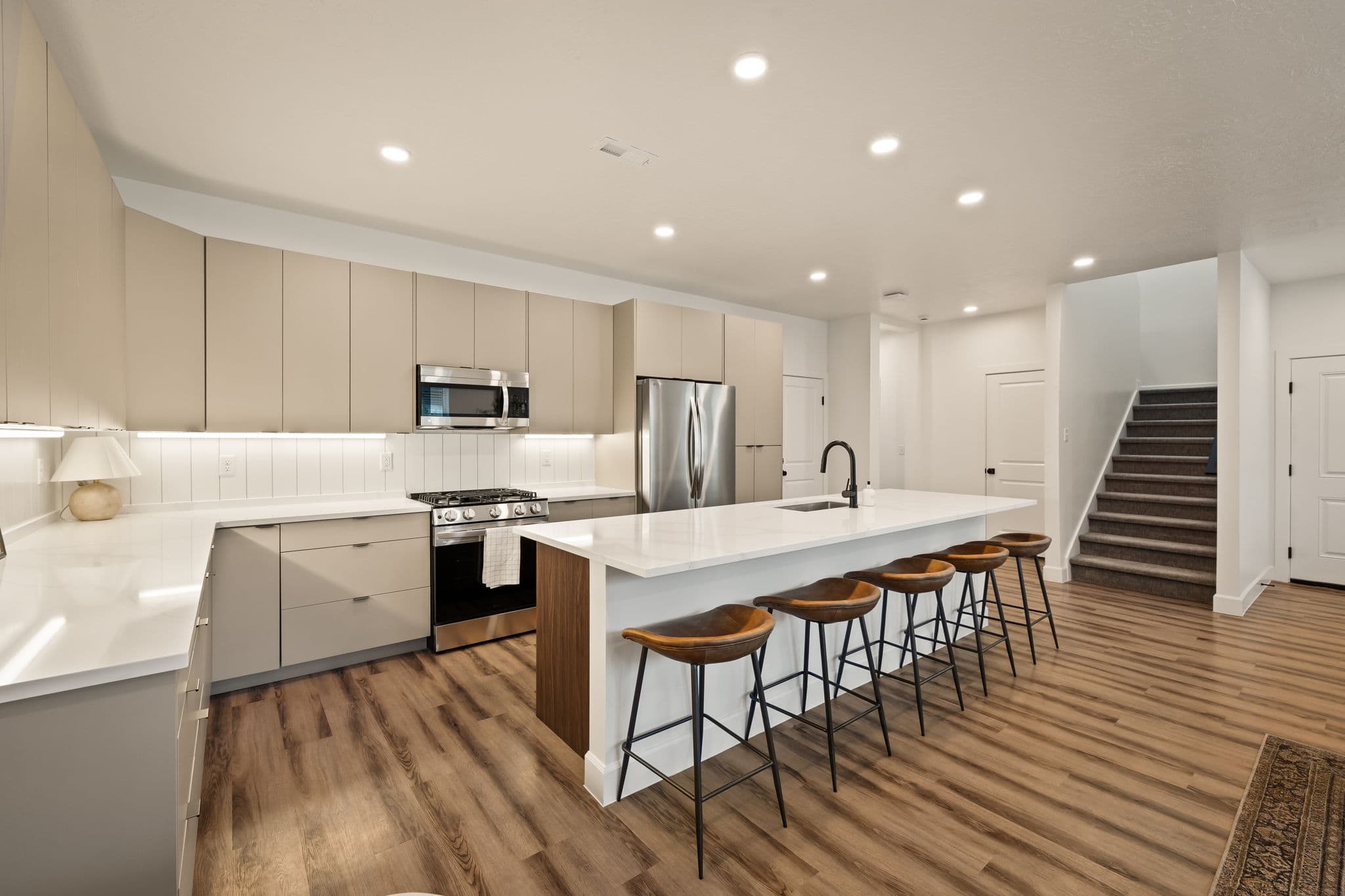 Modern kitchen with light wood cabinetry, stainless steel appliances, a large white island with black faucet, and four wooden bar stools. The space has wood flooring and recessed lighting, with a staircase nearby.