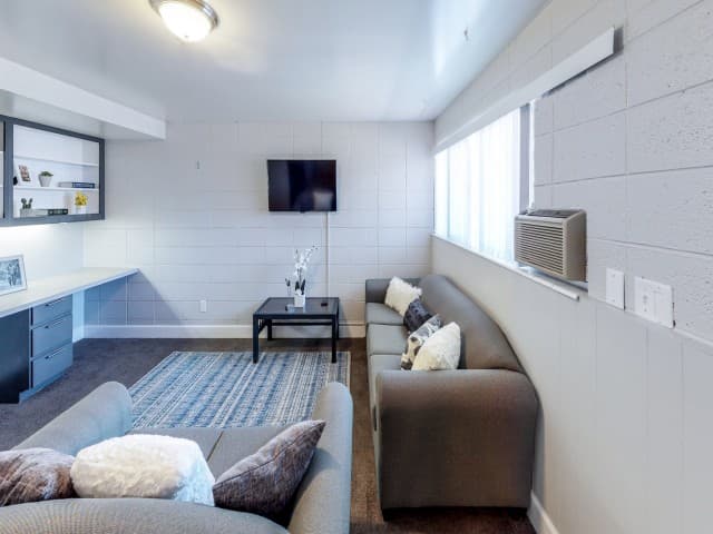 A modern living room at Center Pointe Provo features gray couches, a blue patterned rug, a wall-mounted TV, a black coffee table with a white orchid, a built-in desk with shelves, and a window with blinds and an air conditioner.