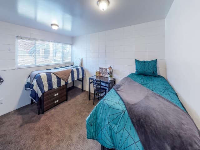 A dorm room at Center Pointe Provo with two single beds, each with colorful bedding, a desk and chair between them, a lamp, and a large window letting in natural light. The room features white walls and a carpeted floor.