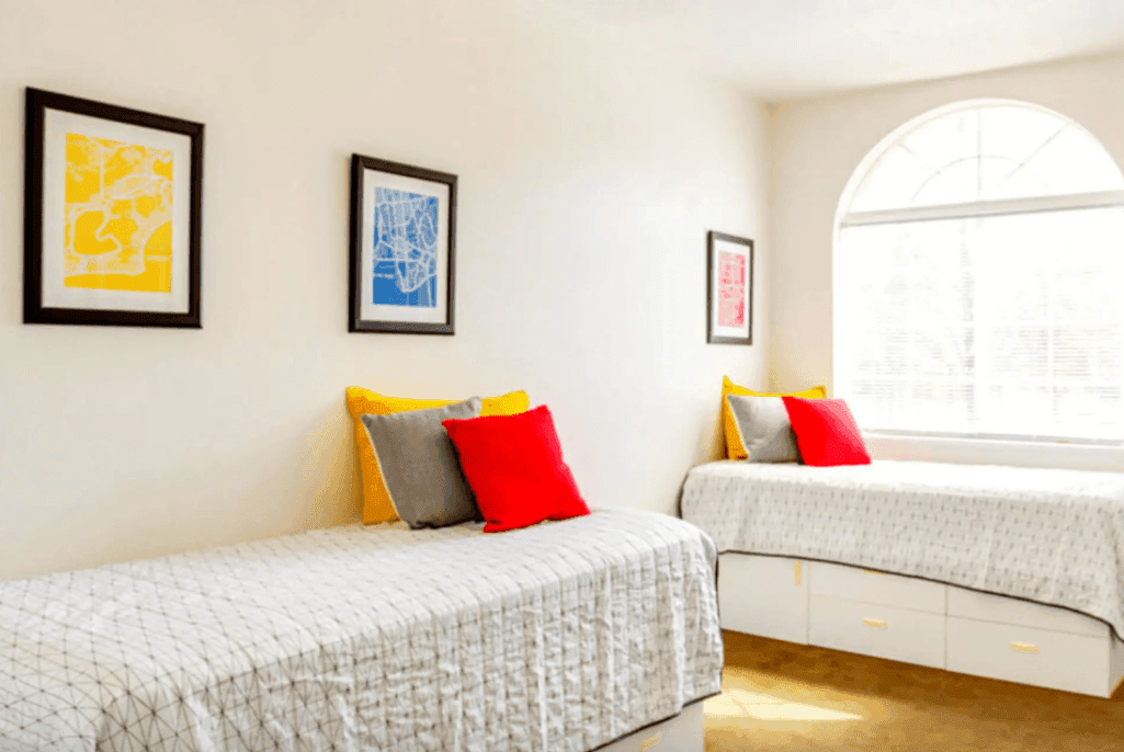 A bright University Towers Orem bedroom with two single beds, patterned gray bedding, yellow and red accent pillows, and drawers underneath. Three framed abstract prints hang on the white wall, and a large arched window lets in sunlight.