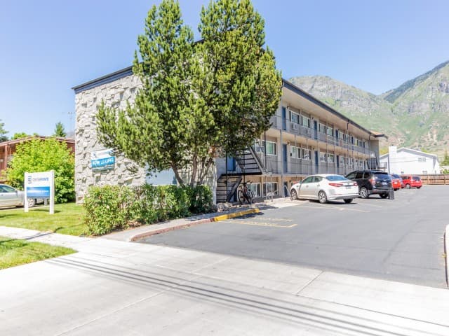 A three-story apartment building with stone and beige exterior at Center Pointe Provo, surrounded by trees and parked cars, with mountains visible in the background on a sunny day.