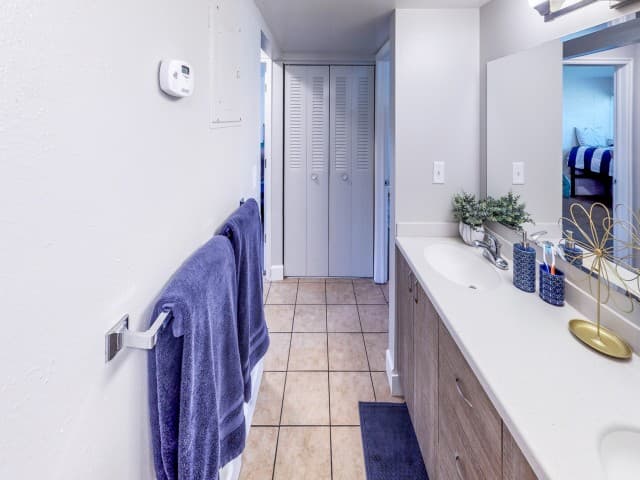 A clean bathroom with beige tile floor, double-sink vanity, white counters, blue towels on the rack, mirror, toiletries, and plants—just steps from the bedroom at Center Pointe Provo. Closet doors are at the end of the room.
