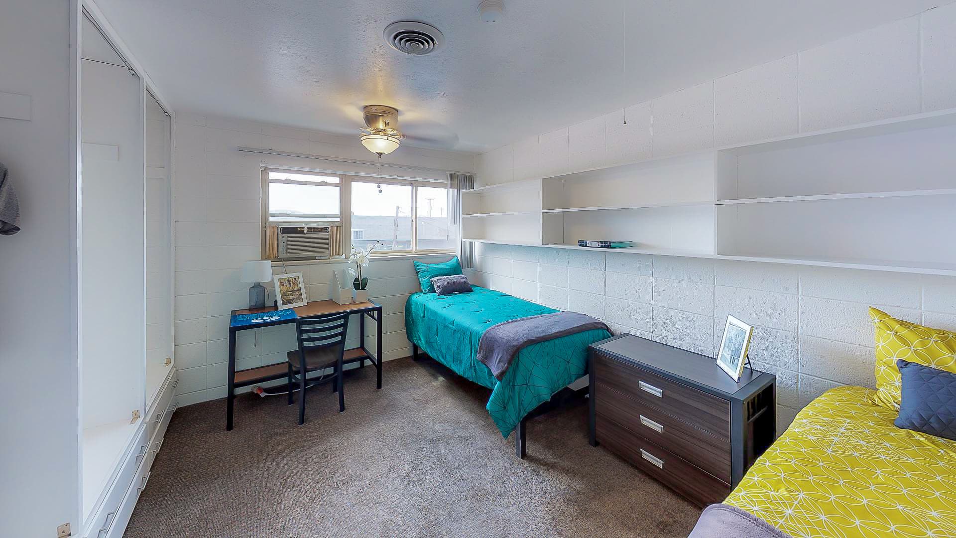 A bright Campus Edge Provo dorm room with two beds—one teal, one yellow—features a desk by the window, wall shelves, and a small dresser between the beds for convenient student living.