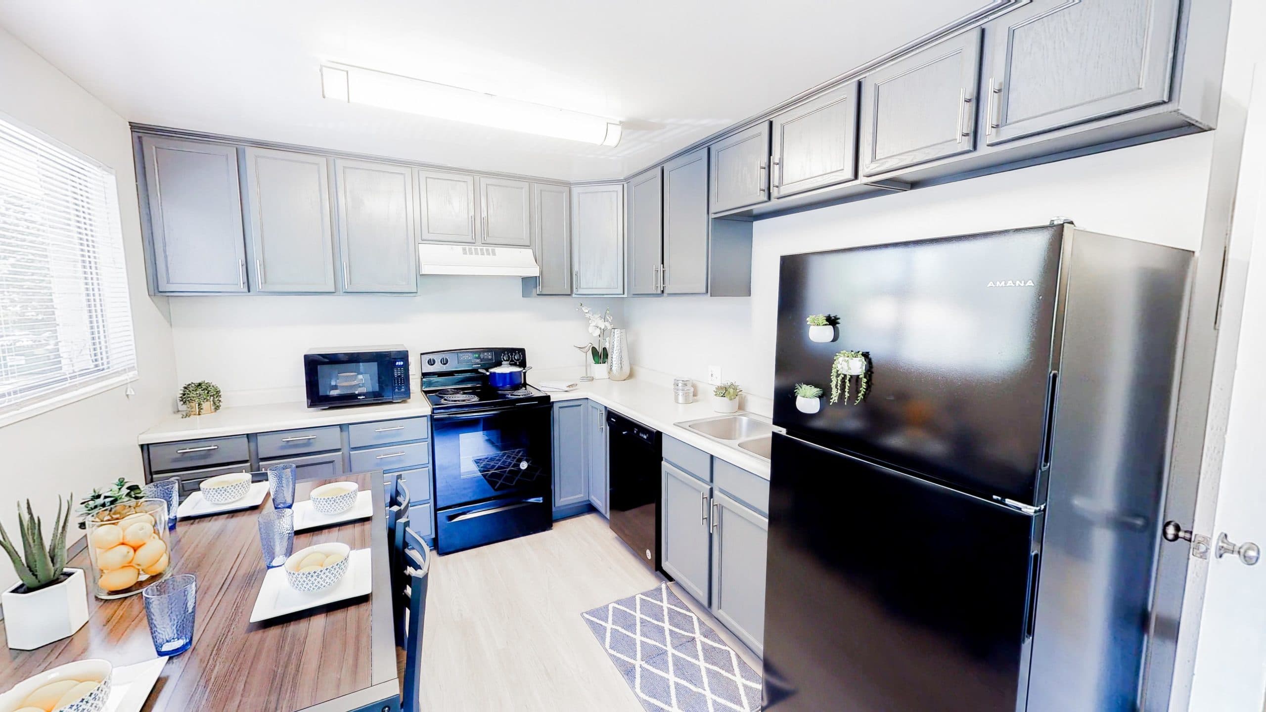 Modern kitchen at Summer Pointe Provo with gray cabinets, black appliances and refrigerator, a dining table set for four with bowls and cups, plants on the counter and fridge, and sunlight streaming in from the left window.