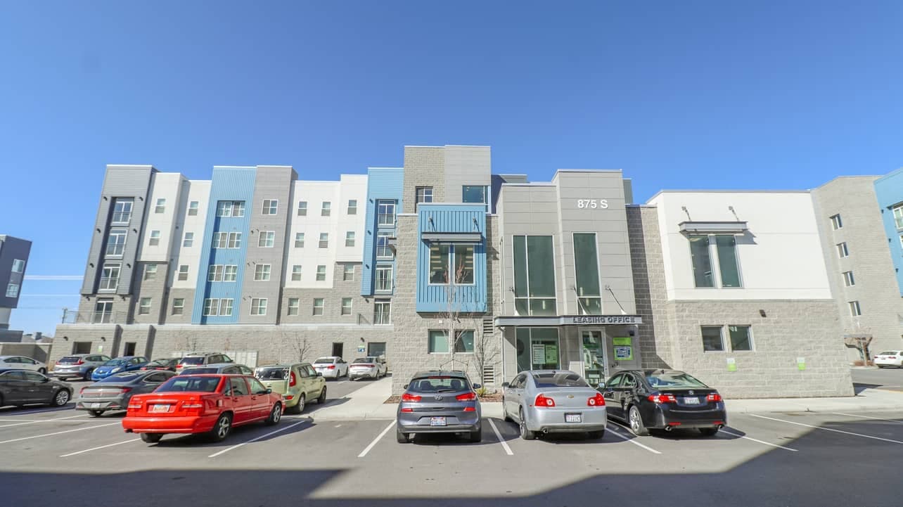 A modern four-story apartment building, Promenade Place Orem features a gray and white exterior, large windows, and a blue balcony. Several cars are parked in front under a clear blue sky. The address 875 S is visible above the entrance.
