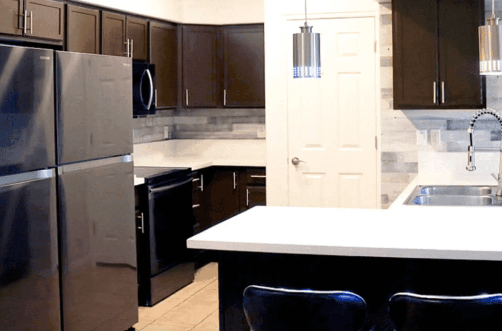 Modern kitchen at University Towers Orem with dark brown cabinets, stainless steel refrigerator and oven, white countertops, double sink, and pendant lights above a breakfast bar with two chairs. Light-colored tile backsplash and floor.