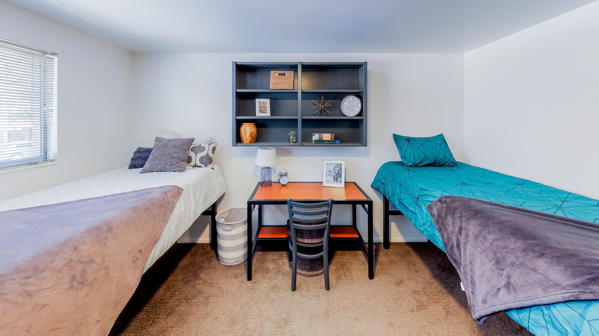 A tidy Summer Pointe Provo dorm room with two single beds, cozy bedding and pillows, a central desk and chair, a lamp, and a dark bookshelf with decor and books mounted above the workspace.