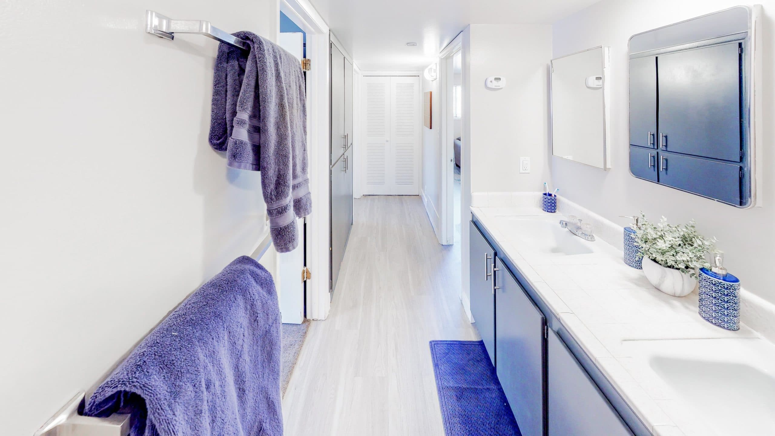 Modern bathroom at Summer Pointe Provo with blue cabinets, white countertops, and blue accents. Two sinks with mirrors, blue towels on a rack, bath mat, and potted plants decorate the space. Hallway with wooden floor beyond.