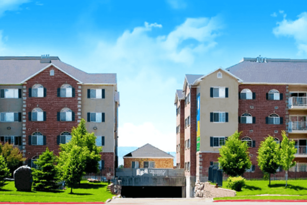 Two multi-story apartment buildings, known as University Towers Orem, feature brick and beige exteriors separated by a driveway to the parking garage. Green trees and grass line the front, with a bright blue sky and light clouds above.