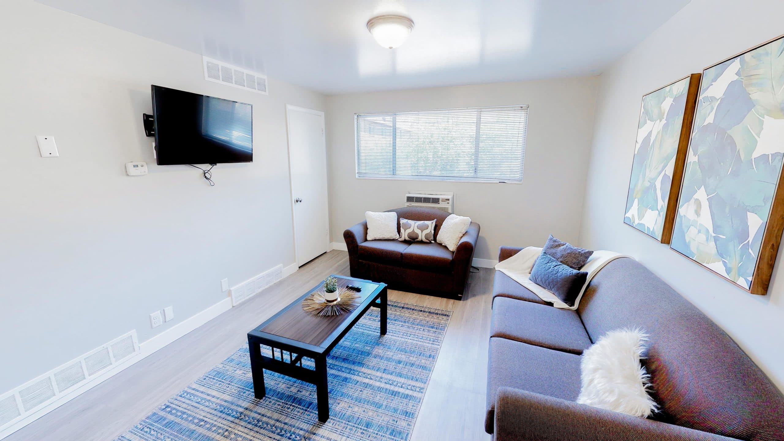A bright living room at Summer Pointe Provo features a brown sofa, matching armchair, coffee table on a blue rug, two large framed artworks, a wall-mounted TV, and a window letting in natural light.