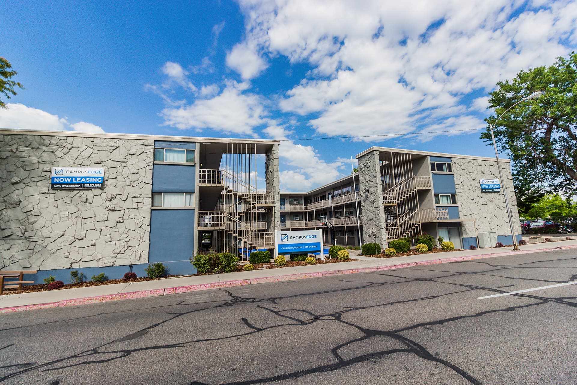 A three-story apartment building with gray stone accents and blue trim, displaying Now Leasing signs on the exterior. The campus edge provo property faces a street with visible pavement cracks under a partly cloudy sky.