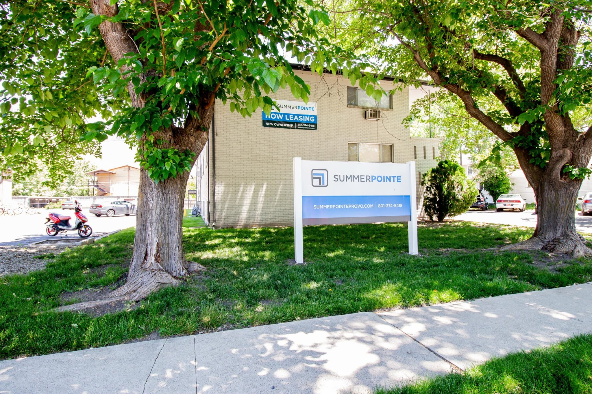 A white sign reading SUMMERPOINTE stands on a grassy lawn between two leafy trees in front of Summer Pointe Provo apartments, featuring a NOW LEASING banner. Cars and a motorcycle are parked nearby.
