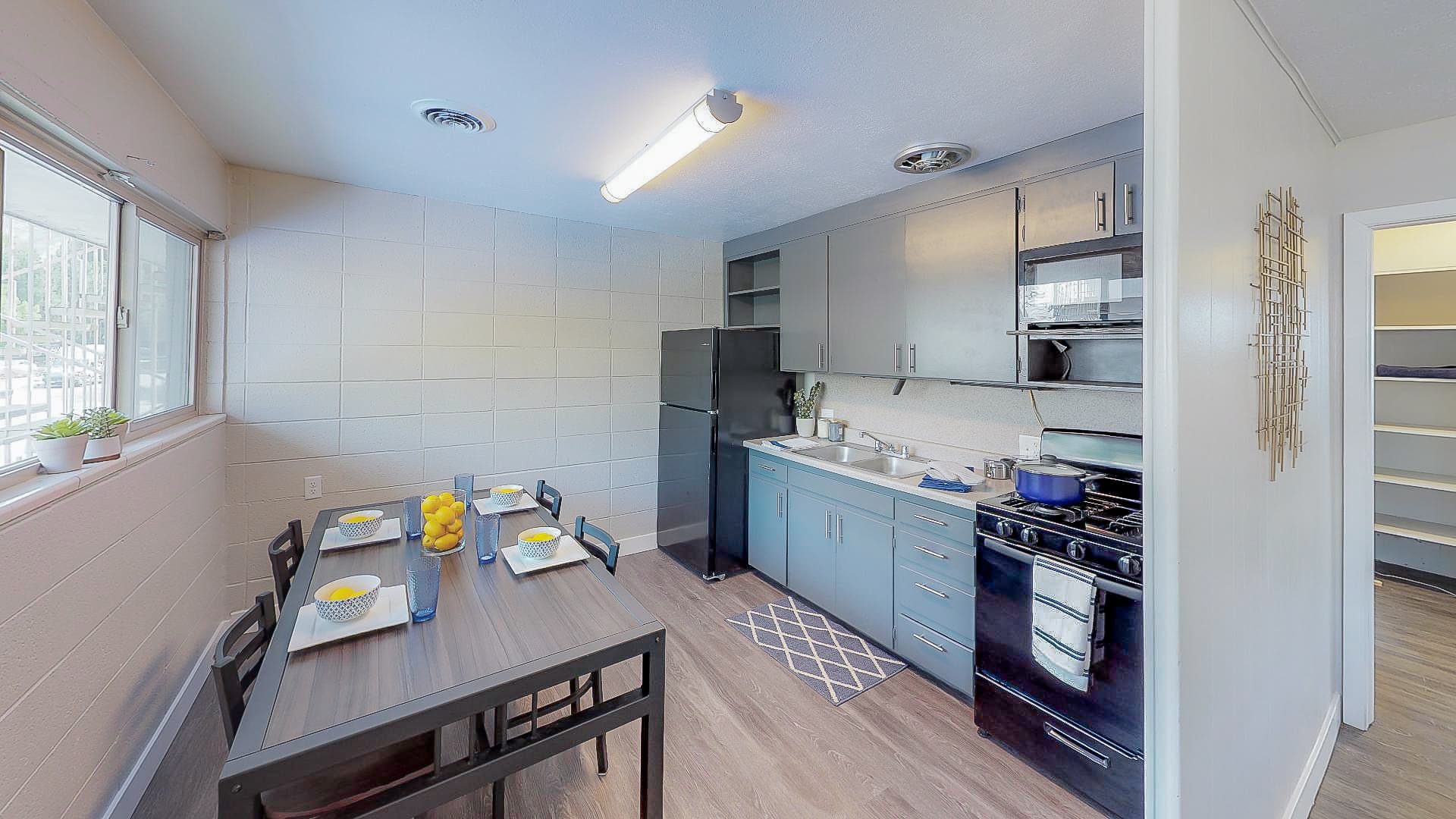 Modern kitchen and dining area at Campus Edge Provo with blue cabinets, black appliances, a set dining table for four, large window, and open shelving. Bright, clean, and organized space with wood-style flooring.