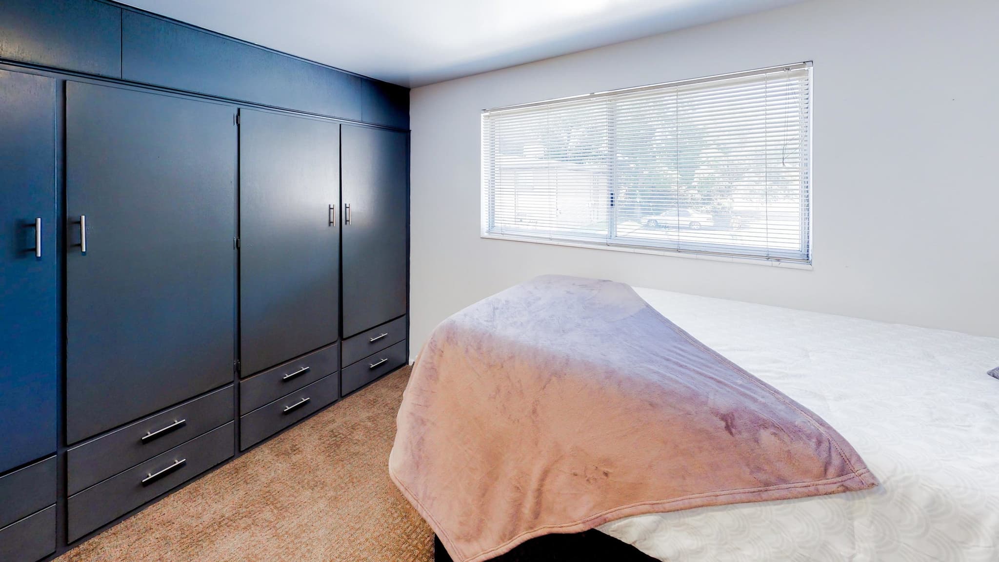A bedroom at Summer Pointe Provo features a large blue built-in wardrobe, a bed with a white quilt and mauve blanket, beige carpet, and a wide window that fills the space with natural light.