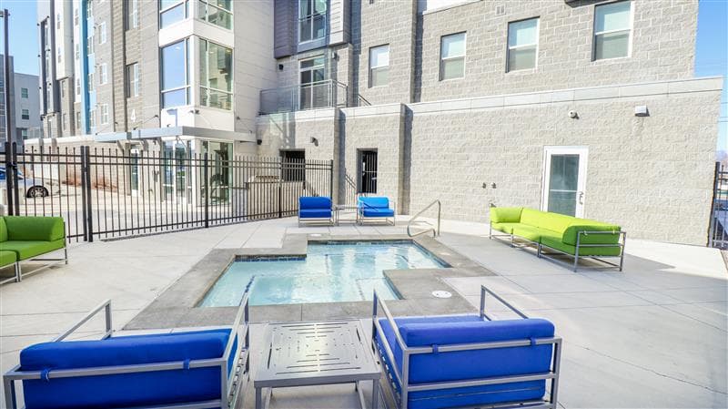 Outdoor courtyard at Promenade Place Orem features a small hot tub surrounded by blue and green cushioned lounge chairs, set beside a modern apartment building with large windows and a gated area.