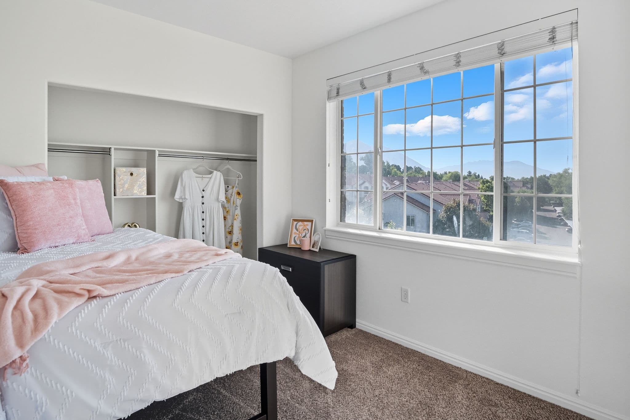 A bright bedroom at Ventana Student Housing Orem features a neatly made bed, an open closet with hanging clothes, a small black dresser, and a large window offering views of clear blue skies and rooftops outside.