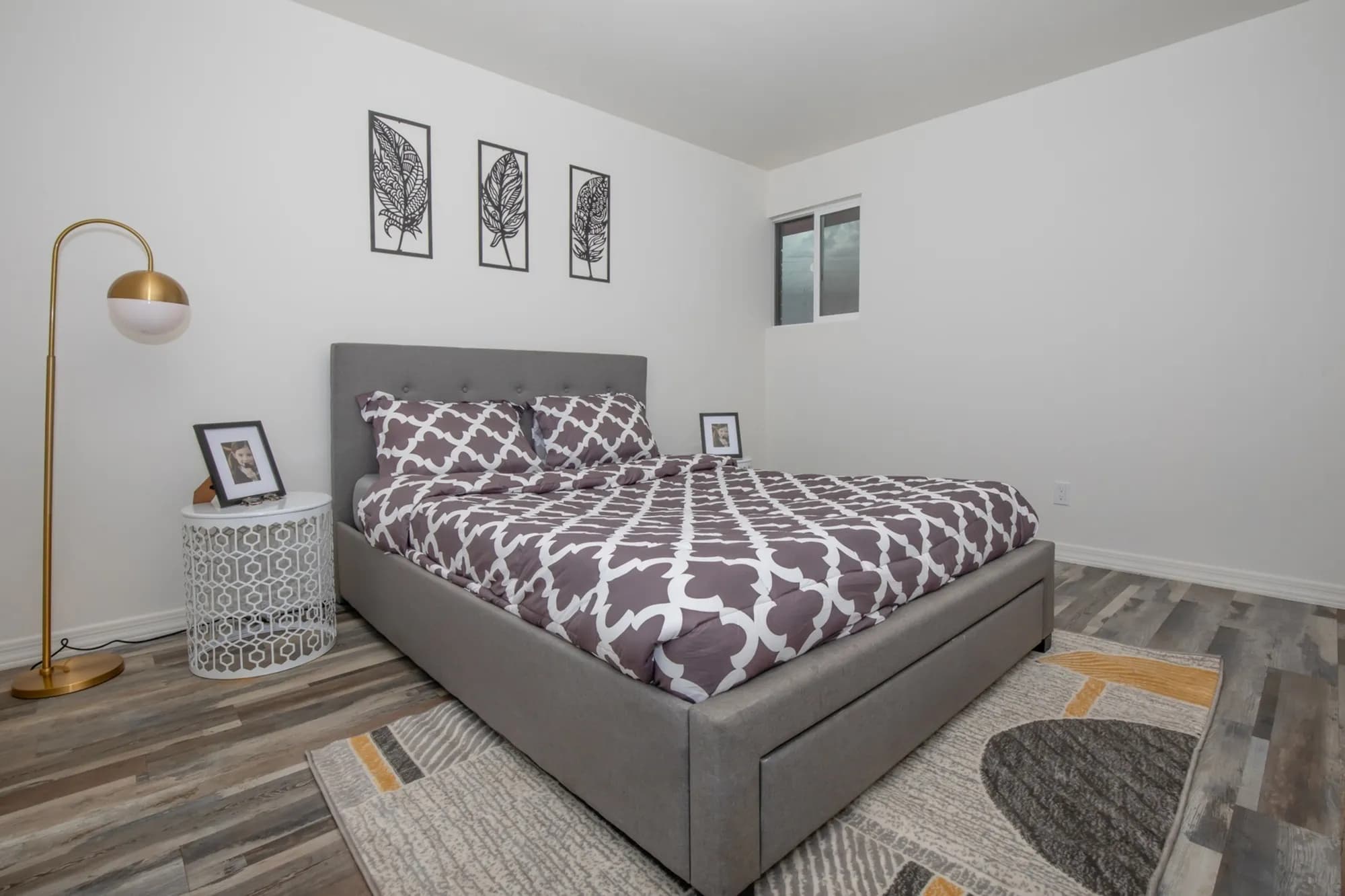 A modern bedroom at Spectra Midtown Apartments Phoenix features a gray upholstered bed, patterned bedding, framed photos on nightstands, a gold floor lamp, three leaf art pieces on the wall, a window, and a patterned rug on wood flooring.