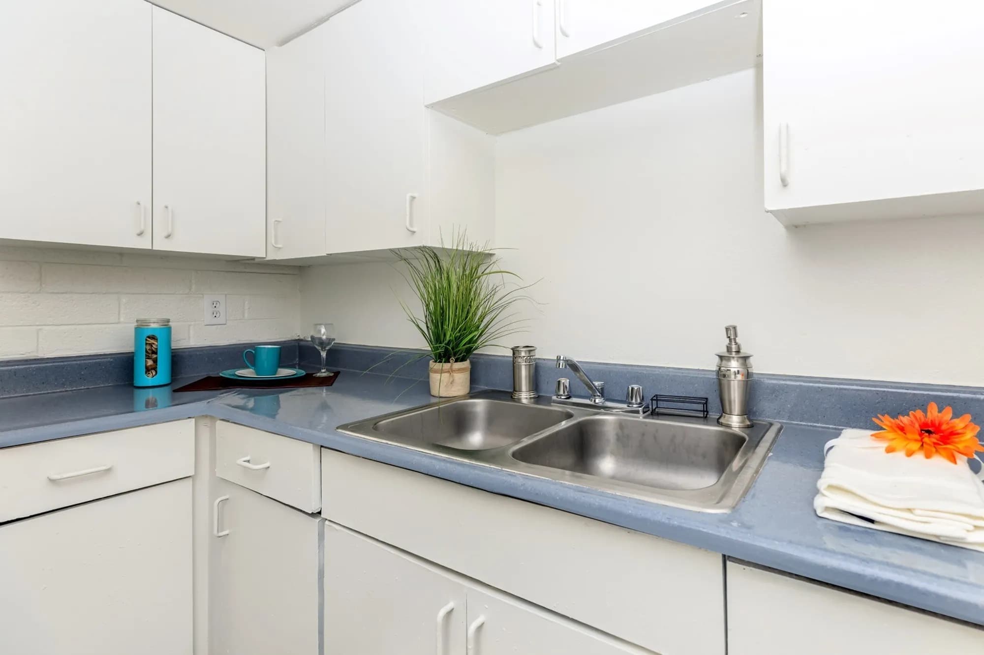 A clean, white kitchen at Spectra Midtown Apartments Phoenix features blue countertops, a stainless steel double sink, white cabinets, a potted plant, a cup with saucer, a candle, and folded towels topped with an orange flower.