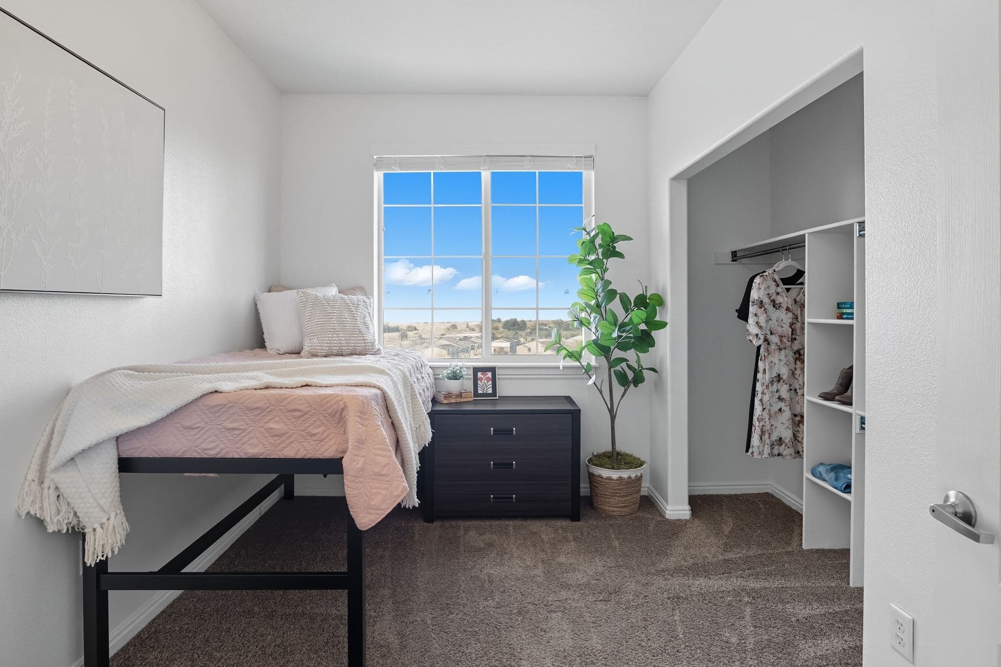 A tidy bedroom at Ventana Student Housing Orem with a raised bed, pink bedding and pillows, a small dresser under a window with a bright sky view, an open closet with clothes, a potted plant, and neutral decor.