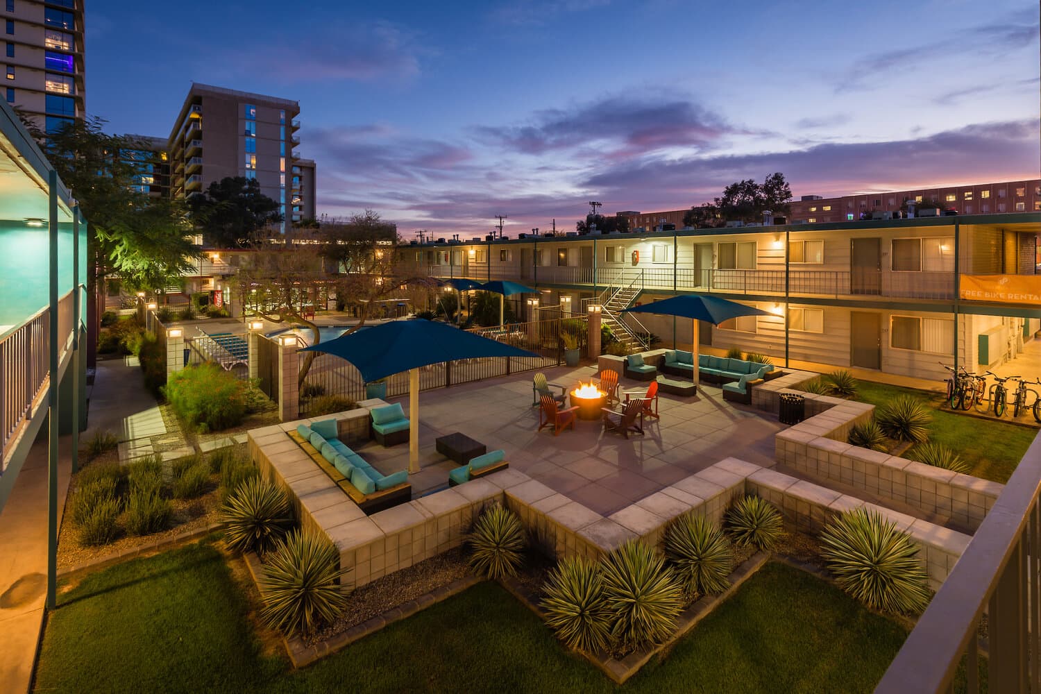 A modern University Pointe Apartments courtyard at dusk with outdoor seating, blue umbrellas, a fire pit, landscaped plants, and surrounding buildings illuminated by soft lights.