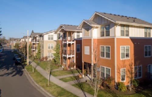 A row of modern, colorful apartment buildings with balconies lines 7th Street Station, a quiet residential street under a clear blue sky. Leafless trees and neatly trimmed grass border the sidewalk.