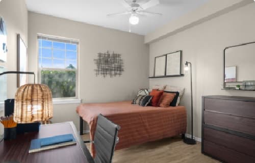 A tidy bedroom at 7th Street Station features a bed with rust-colored bedding, a desk with a chair and lamp, a dresser, wall art, and a window letting in natural light. The room has light walls and wooden flooring.