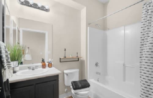 Modern bathroom at 7th Street Station with a white bathtub and striped shower curtain, toilet with folded towels on top, sink with soap dispensers, mirror, wall shelf, and a potted plant on the counter.
