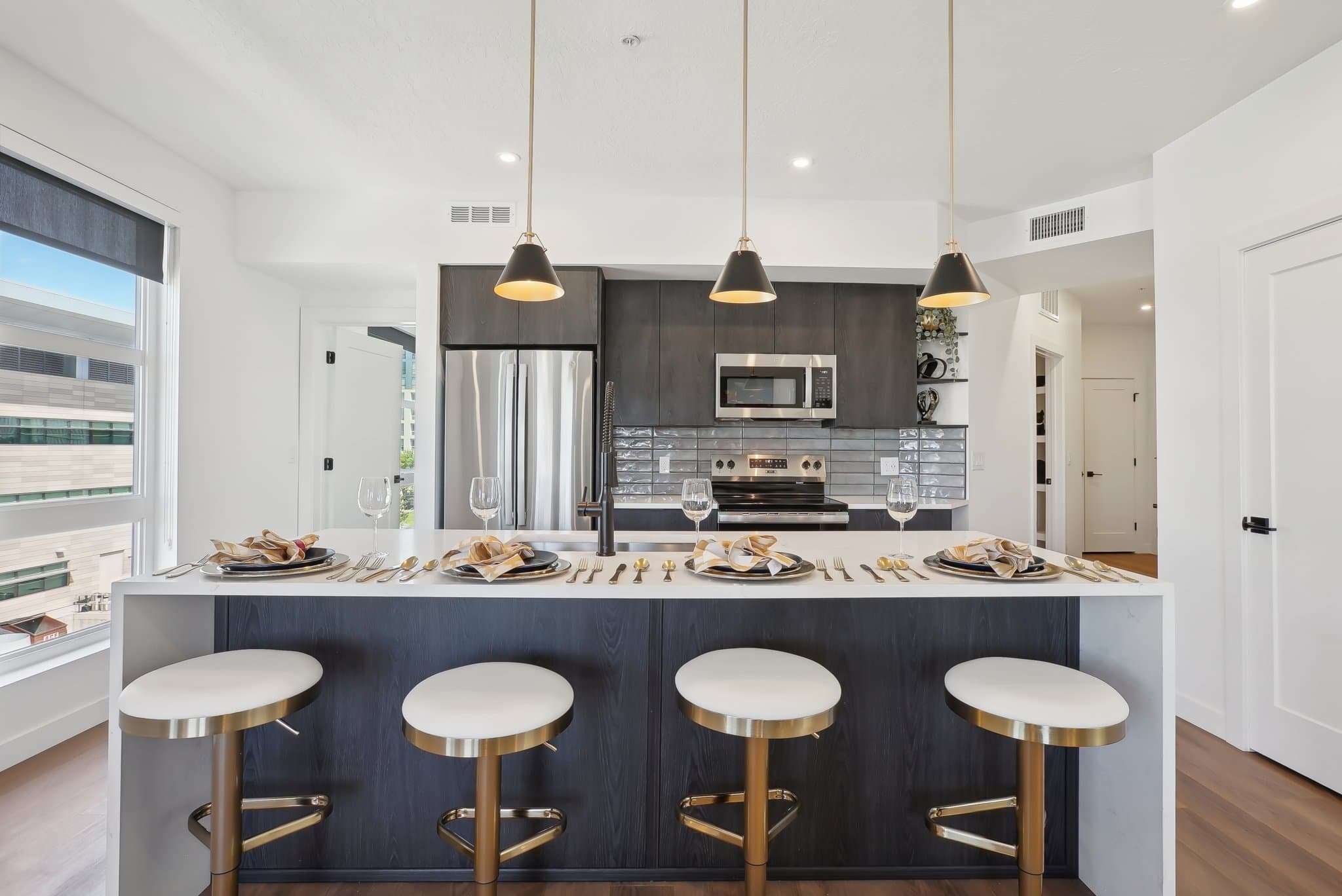 Modern kitchen with a white Liberty Duet island, four gold and white barstools, pendant lights, dark cabinets, stainless steel appliances, and place settings on the counter, with large windows bringing in natural light.