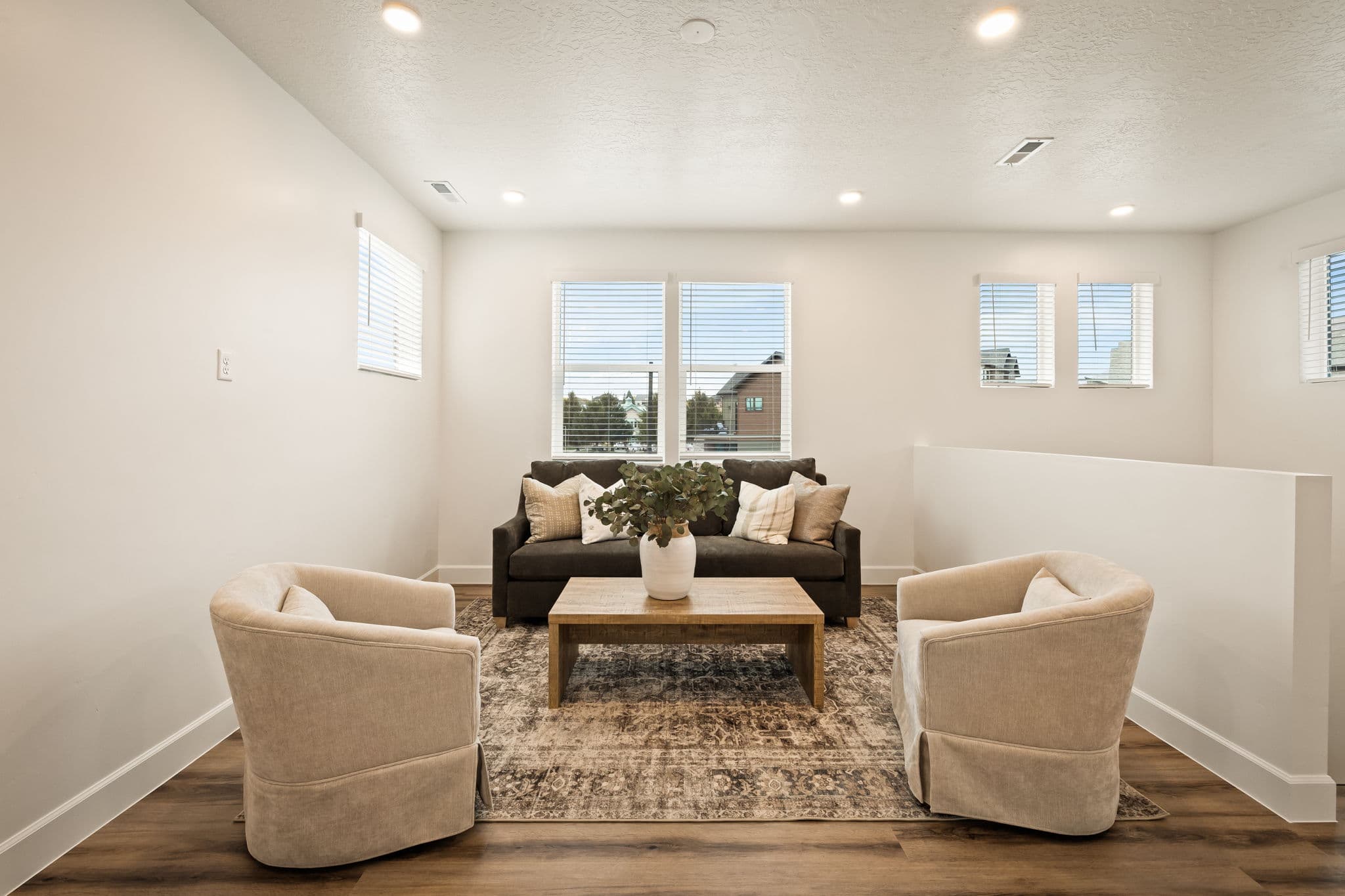 A bright, modern living room with a dark sofa, two beige armchairs, a wooden coffee table with a potted plant, and large windows letting in natural light. The space features light walls and hardwood flooring.