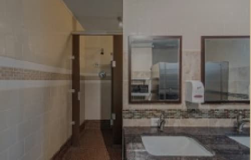 A public restroom at 909 14th Boulder features a brown stall door on the left, two sinks with soap dispensers below two mirrors on the right, and tiled walls and countertops throughout.