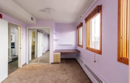 A small, empty room at 909 14th Boulder with light purple walls, brown carpet, two large windows with wooden frames, a built-in desk, a radiator under the windows, and mirrored closet doors.
