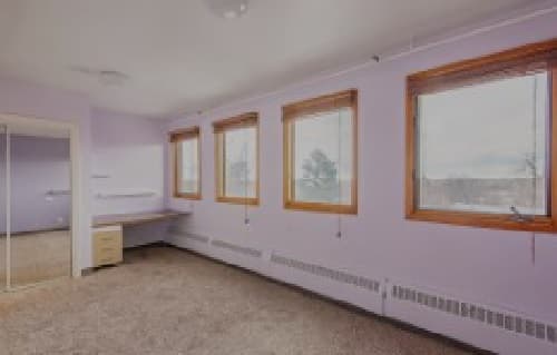 A spacious, empty room at 909 14th Boulder features light purple walls, three large windows with wooden frames, a built-in desk, and a mirrored closet. The carpeted floor and ample natural light make the space bright and inviting.