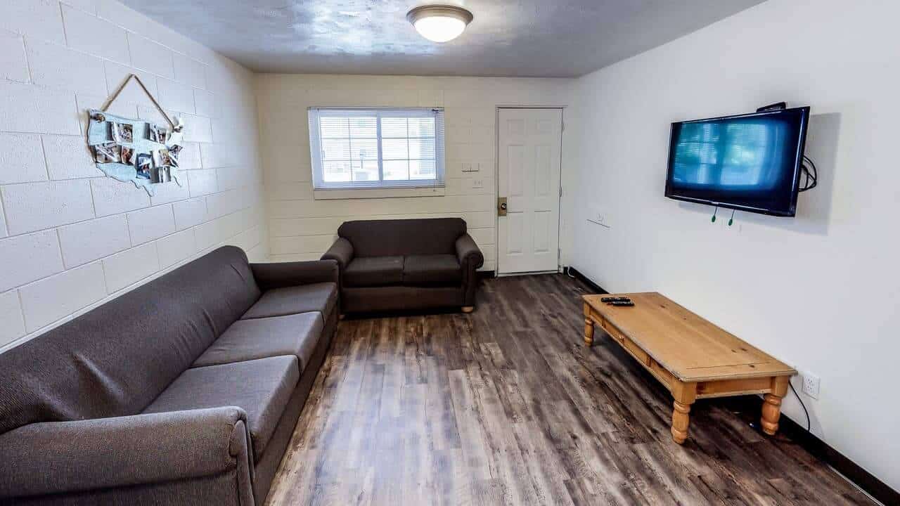 A small living room in Alpine Chalet Apartments Rexburg features gray couches, a wooden coffee table, a wall-mounted TV, and a sunlit window. Photos decorate the wall, while dark wood floors add warmth to the tidy, simply furnished space.
