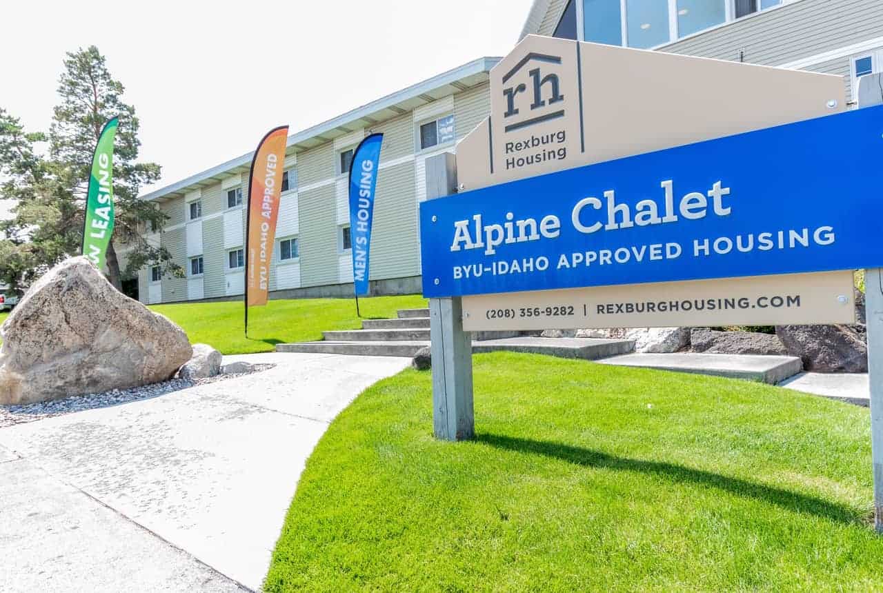 A blue sign for Alpine Chalet Apartments Rexburg, BYU-Idaho approved housing by Rexburg Housing, stands on a lawn with colorful flags and a white apartment building in the background.